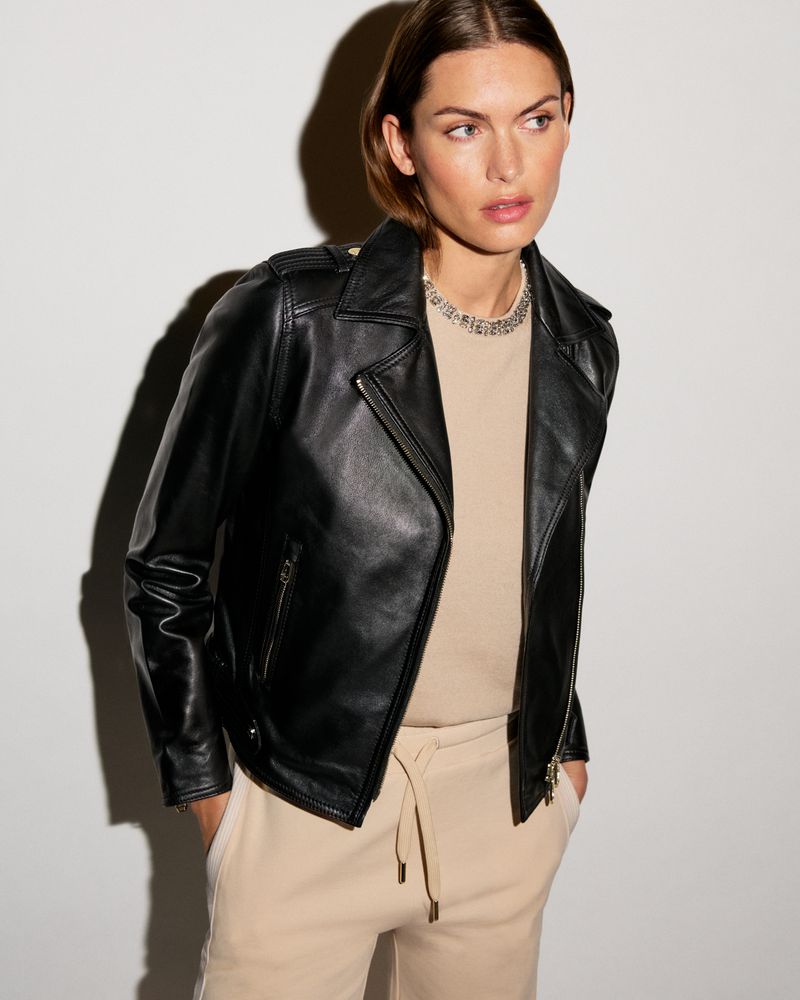 Adult woman in a black leather jacket and beige pants stands in a photo studio setting against a neutral background. Beauty and fashion.