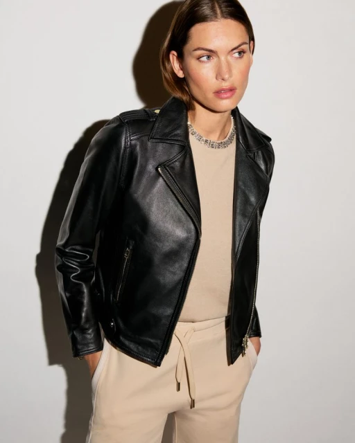 Adult woman in a black leather jacket and beige pants stands in a photo studio setting against a neutral background. Beauty and fashion.