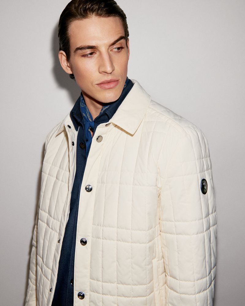 Portrait of a young adult male wearing a cream-colored quilted jacket over a blue collared shirt, posing in front of a neutral background.
