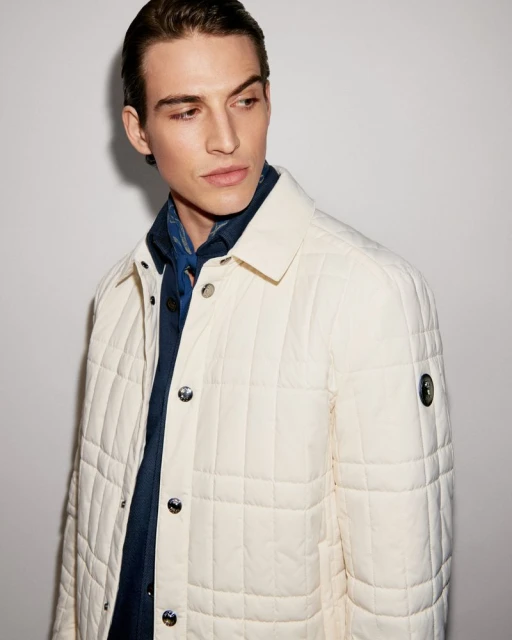 Portrait of a young adult male wearing a cream-colored quilted jacket over a blue collared shirt, posing in front of a neutral background.