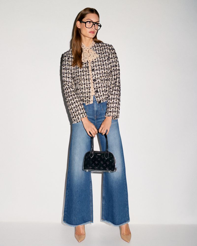 A stylish woman with glasses, a tweed jacket, wide-leg jeans, and nude heels holds a small black handbag against a neutral backdrop.