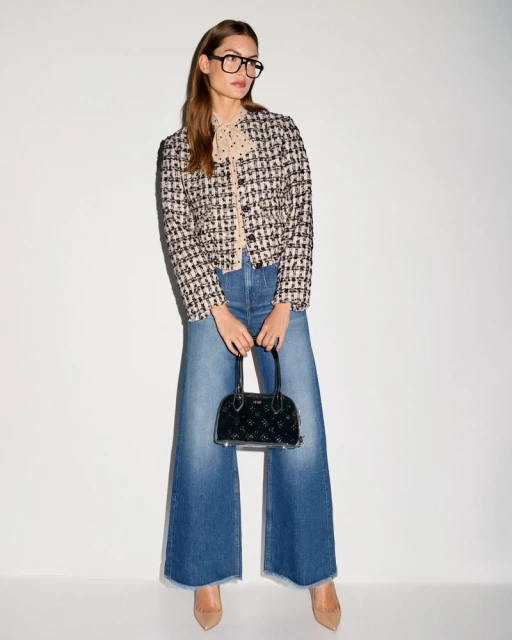 A stylish woman with glasses, a tweed jacket, wide-leg jeans, and nude heels holds a small black handbag against a neutral backdrop.