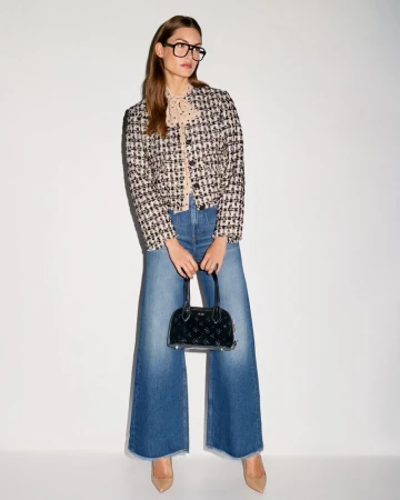 A stylish woman with glasses, a tweed jacket, wide-leg jeans, and nude heels holds a small black handbag against a neutral backdrop.