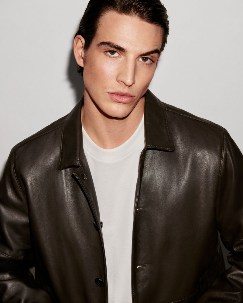 A portrait of a handsome man wearing a brown leather jacket over a white t-shirt, set against a neutral background in a studio shot.