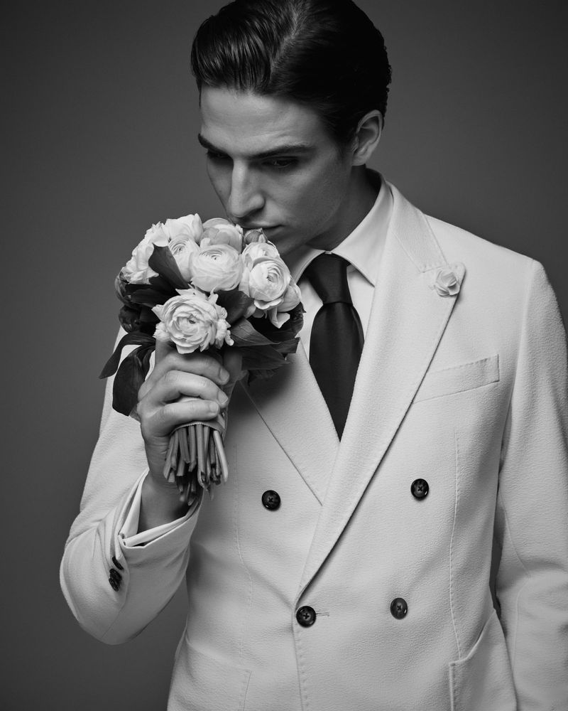 Black and white portrait of a man in a suit holding a bouquet of flowers, close to his face, in a studio setting with a grey background.