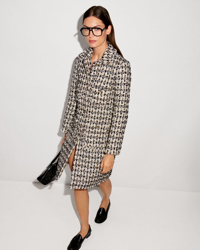 Adult female wearing a coat dress, black glasses, and loafers standing in front of a white background in a fashion studio shot.