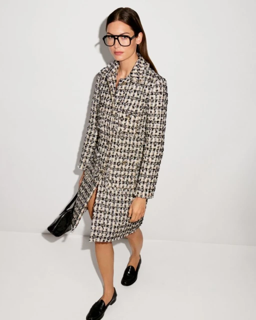 Adult female wearing a coat dress, black glasses, and loafers standing in front of a white background in a fashion studio shot.