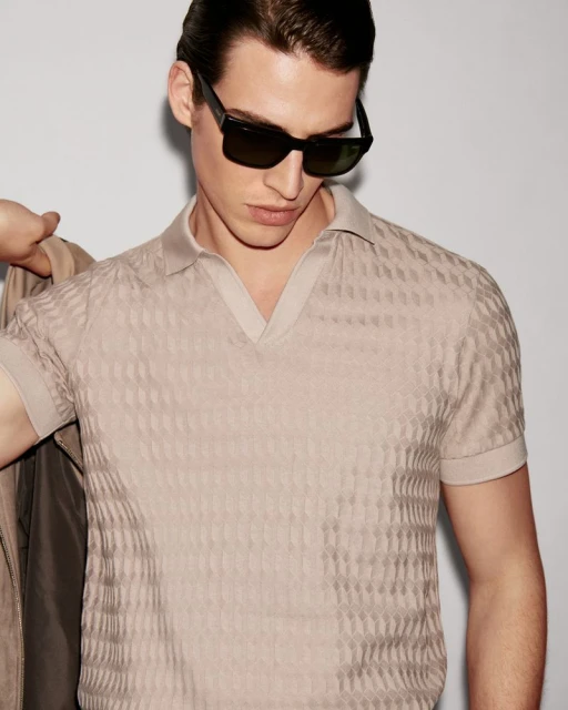 Close-up of a man in a textured beige polo shirt and sunglasses, holding a jacket; stylish, modern studio shot.