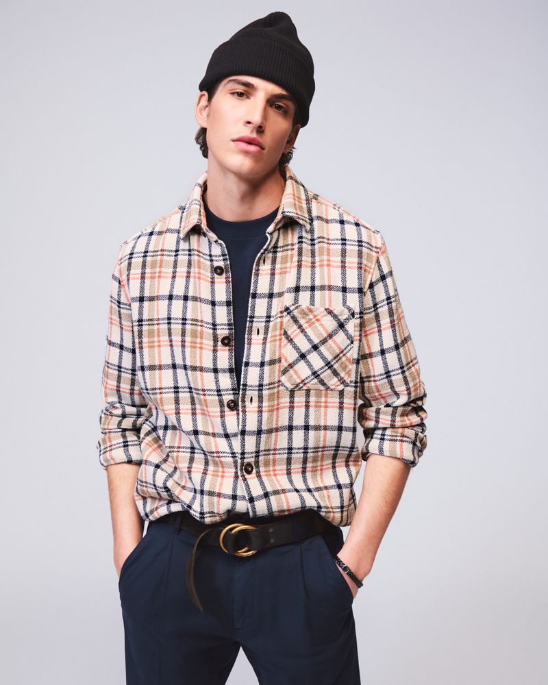 Portrait of a young adult male wearing a flannel shirt, beanie hat, and dark trousers in a studio setting with soft lighting.