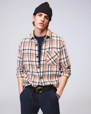Portrait of a young adult male wearing a flannel shirt, beanie hat, and dark trousers in a studio setting with soft lighting.