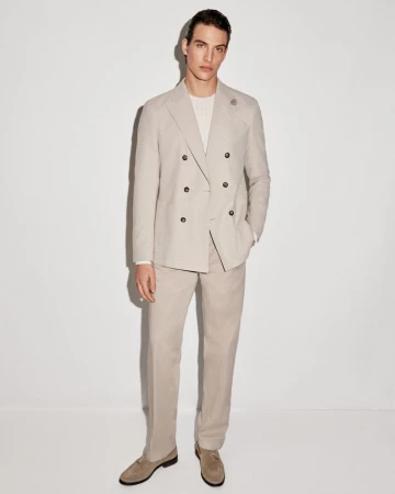Model Amerigo Valenti in a light beige double-breasted suit and suede loafers standing against a plain white studio background.