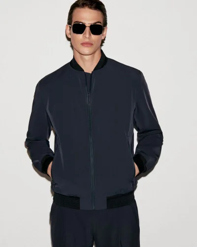 Amerigo Valenti in dark sunglasses and a navy zip-up bomber jacket against a light studio backdrop.