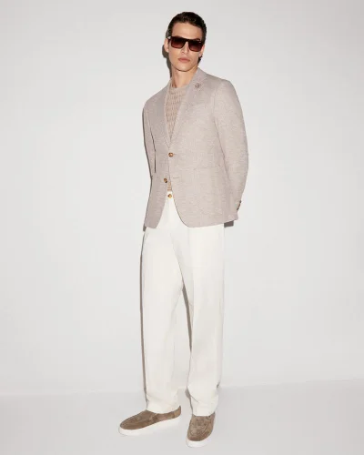 Model wearing a light beige blazer, textured sweater, and white trousers against a plain white studio backdrop.