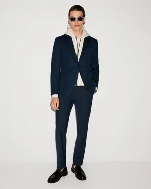 Male model Amerigo Valenti wearing a navy blue suit over a cream hoodie with sunglasses against a white studio background.