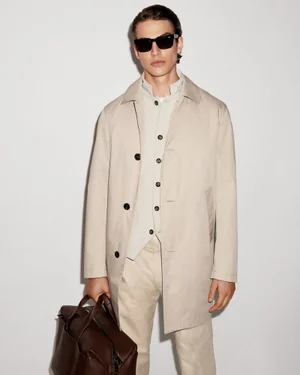 Male model in a beige coat and sunglasses holding a dark brown leather bag against a white seamless background