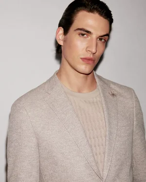 Portrait of a male model wearing a textured beige blazer and crewneck sweater against a plain light grey background.