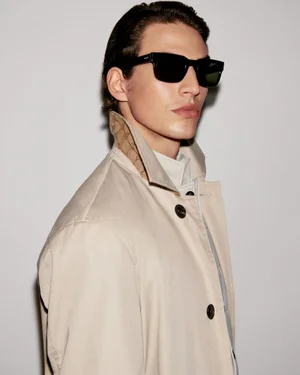 Model wearing dark sunglasses and a light beige trench coat against a plain neutral background.
