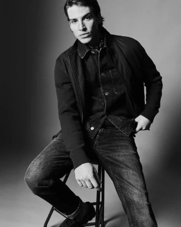 Model Amerigo Valenti seated on a stool wearing a dark jacket, denim shirt, and jeans in a black and white studio portrait.