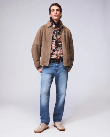 Full-length portrait of a male model wearing a brown suede jacket, patterned shirt, and blue jeans against a neutral studio background