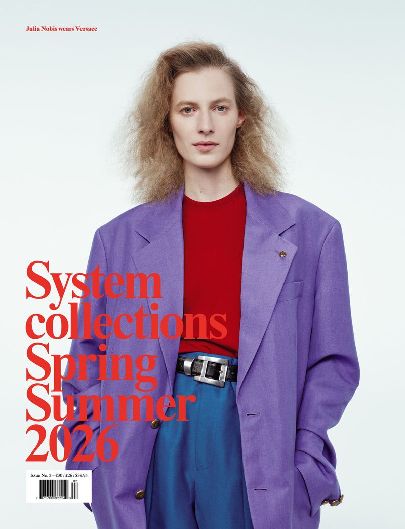 Editorial fashion shot of a blonde adult female wearing a purple blazer, red shirt, and blue pants against a bright white backdrop.