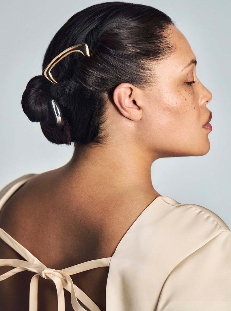 Profile view of an adult female with dark hair styled in a bun with two gold hair accessories. The woman is wearing a cream-colored dress.