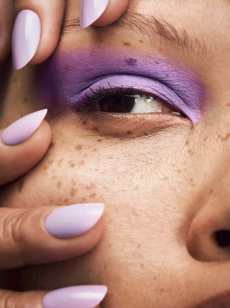 Close-up of a face with freckles, lavender eyeshadow, and light purple stiletto nails framing the face, showcasing beauty and makeup.