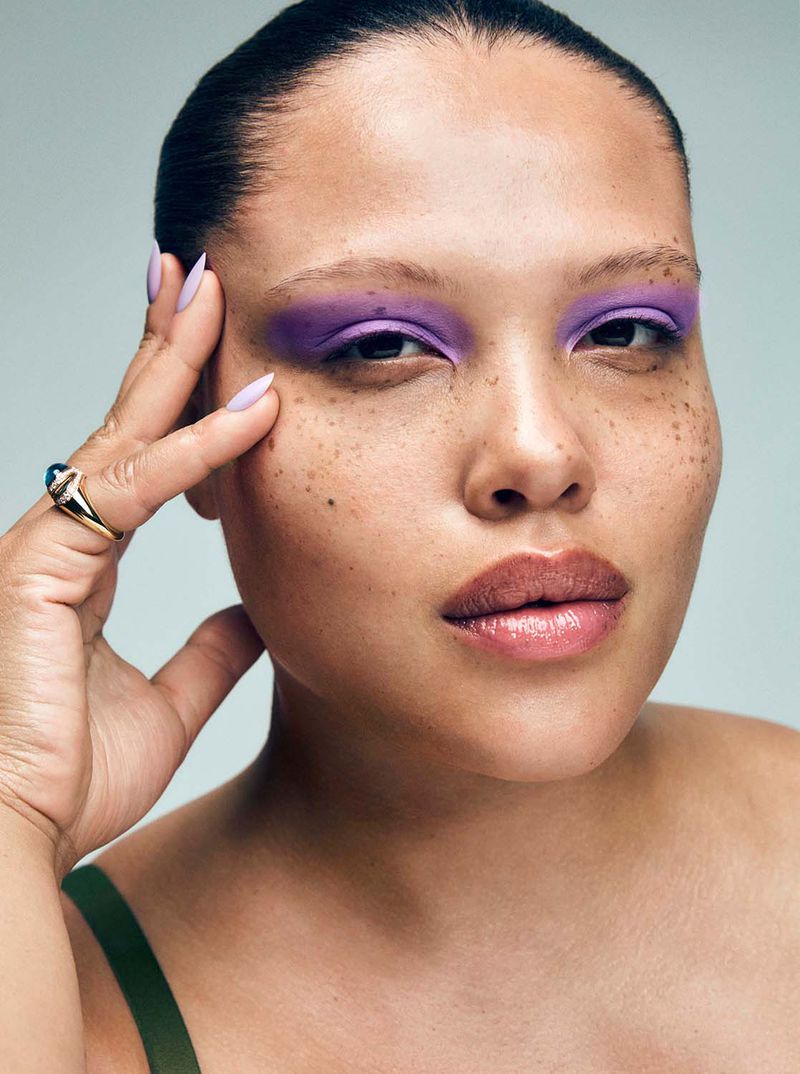 Close-up portrait of an adult woman with freckles wearing purple eyeshadow, gold ring, and lilac nail polish against a light background.