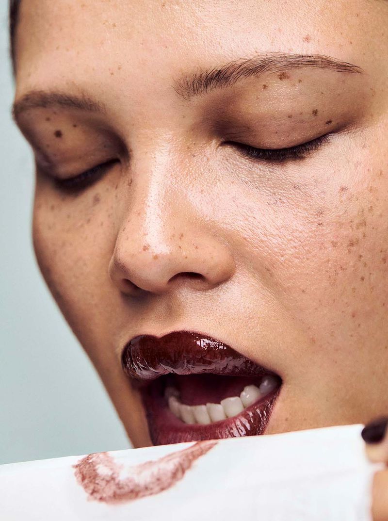 Close-up shot of a woman with closed eyes and freckles, wearing dark lipstick and blotting her lips with a white cloth showing a lipstick print.