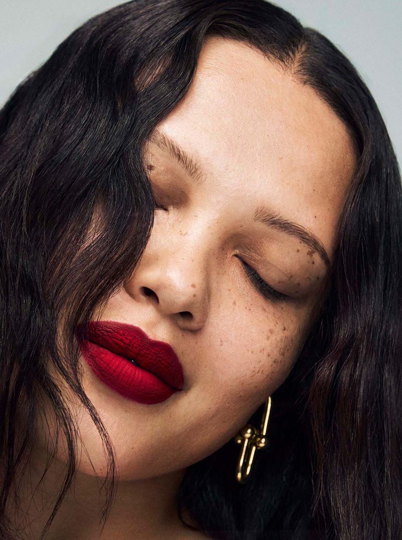 Portrait of a woman with dark wavy hair and freckles, wearing dark red lipstick, with her eyes closed and a gold earring.