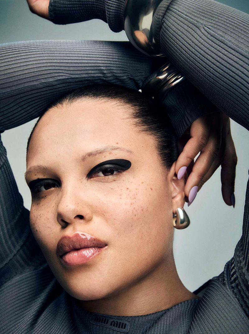 Close-up portrait of a woman with black eyeshadow and glossy lips wearing a grey ribbed top and silver jewelry in a studio setting.