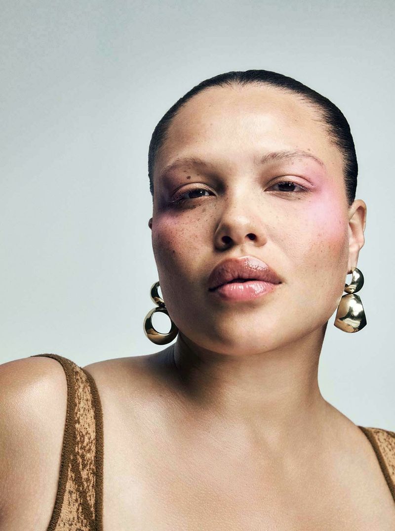 Close-up portrait of a woman with pink eye shadow, gold earrings, and a neutral-toned top against a light grey background.
