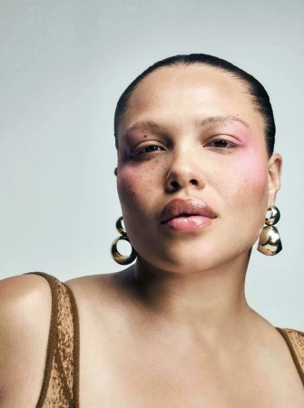 Close-up portrait of a woman with pink eye shadow, gold earrings, and a neutral-toned top against a light grey background.