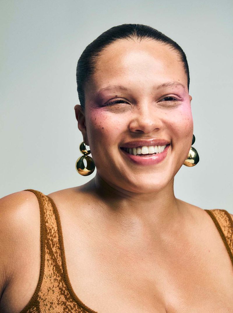 Smiling woman with pink eyeshadow and gold ball earrings posing in a photo studio with neutral background and soft lighting.