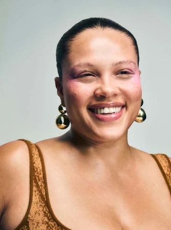 Smiling woman with pink eyeshadow and gold ball earrings posing in a photo studio with neutral background and soft lighting.
