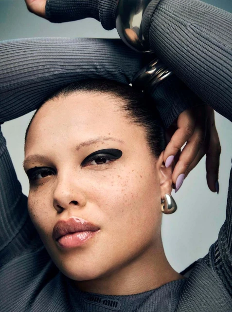 Close-up portrait of model Alva Claire wearing a ribbed grey top with arms raised behind her head, featuring bold black eyeliner.