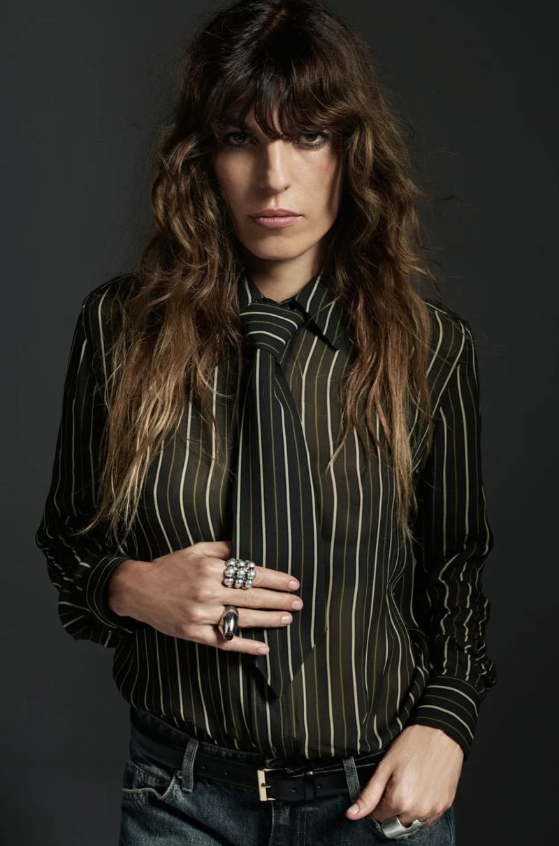A woman with long brown hair wears a striped blouse and jeans against a dark gray background in a studio fashion shot.