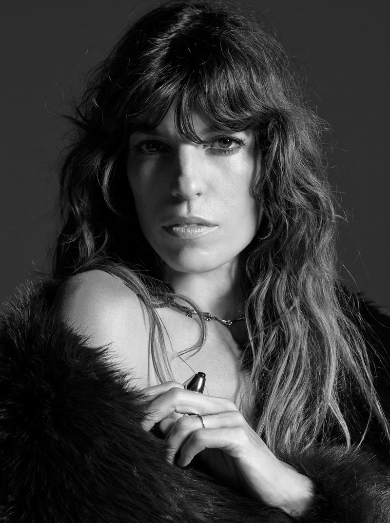 Black-and-white portrait of an adult woman with wavy hair and a serious expression, wearing a fur coat and a delicate necklace indoors.