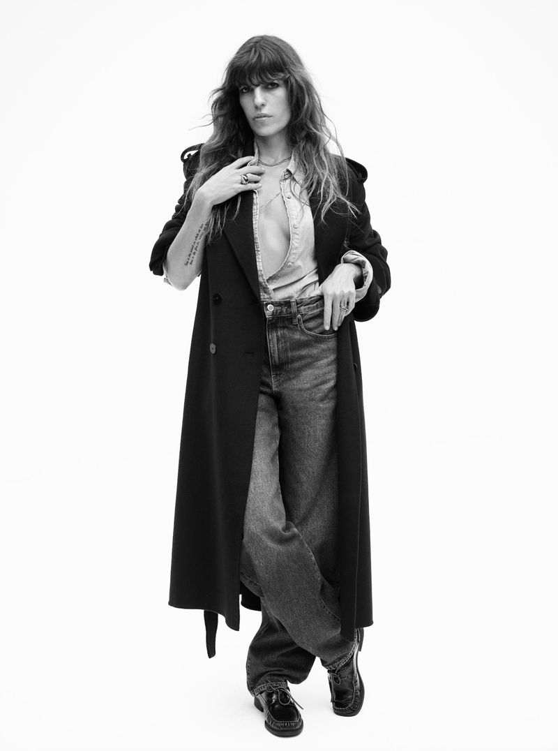 Full-length portrait of a woman in a dark overcoat and denim clothing, posing against a white background in a fashion editorial.