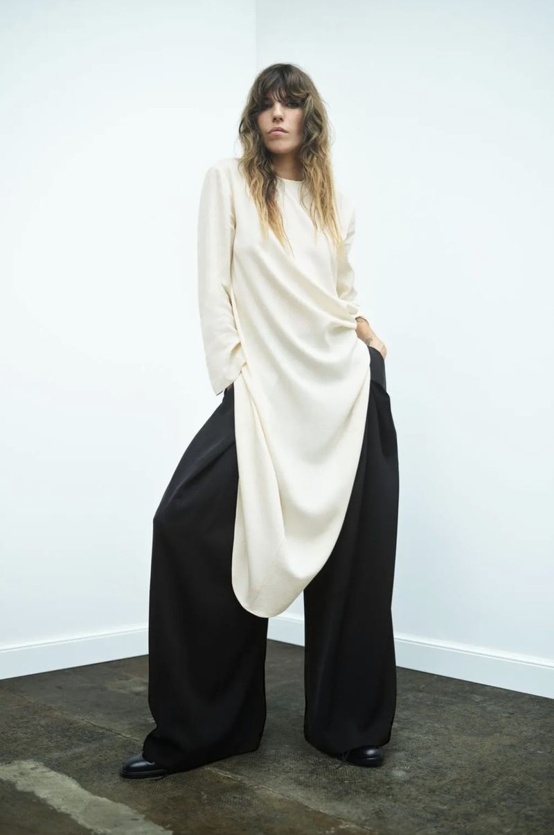 Lou Doillon wearing an asymmetrical cream draped longline tunic and wide-leg black trousers against a white minimalist corner.