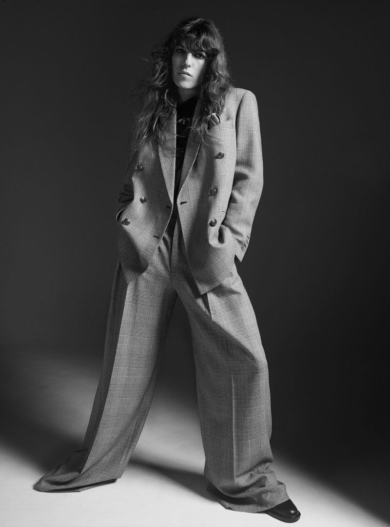 Adult woman with long brown hair poses in a gray double-breasted suit with wide leg pants against a neutral studio backdrop.