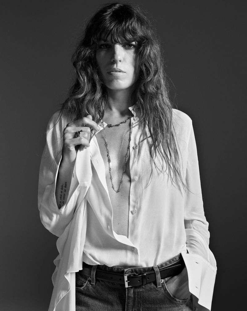 Monochrome portrait of Lou Doillon wearing an unbuttoned white shirt and denim jeans against a dark studio background.