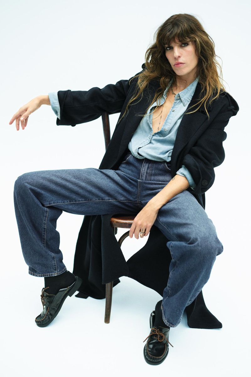 A brunette woman with bangs sits on a wooden chair, wearing a dark coat, light blue shirt, blue jeans, and black shoes against a white backdrop.