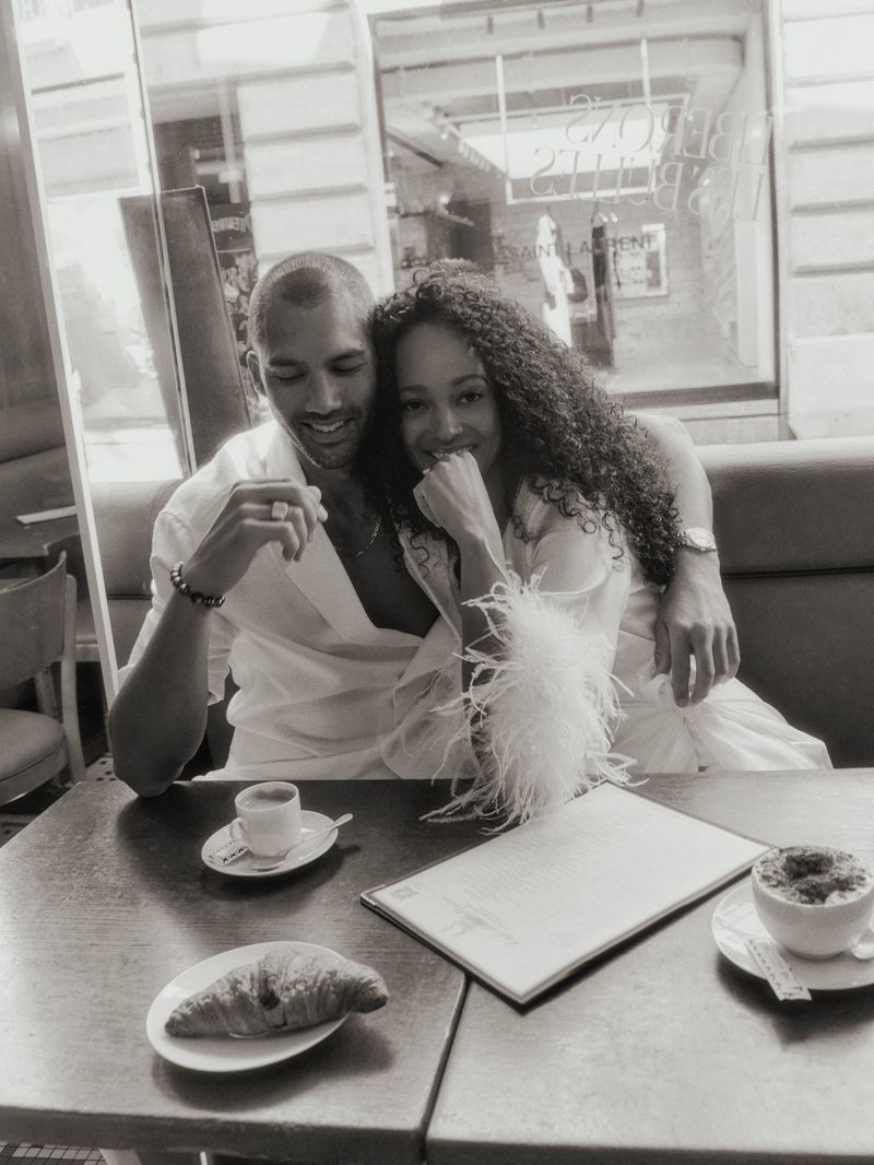 Romantic black and white photo of a couple at a cafe table with coffee and croissant. The light is diffused and serene.