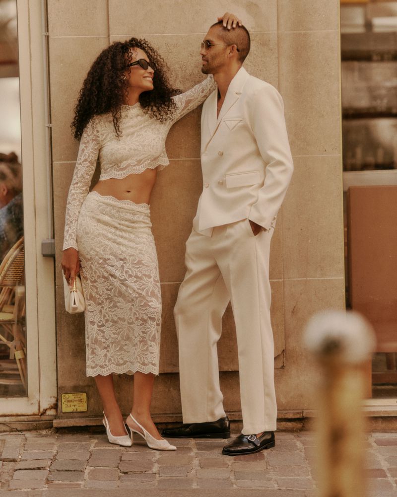 Stylish couple posing in coordinated ivory outfits outside, woman in lace skirt and crop top, man in double-breasted suit, sunglasses.