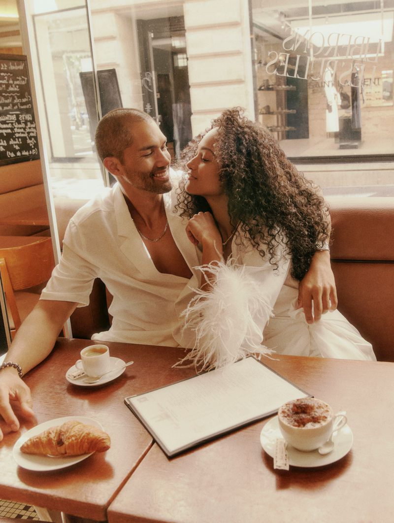 A couple dressed in white sit close together at a cafe table with coffee and a croissant, creating an intimate, romantic atmosphere.