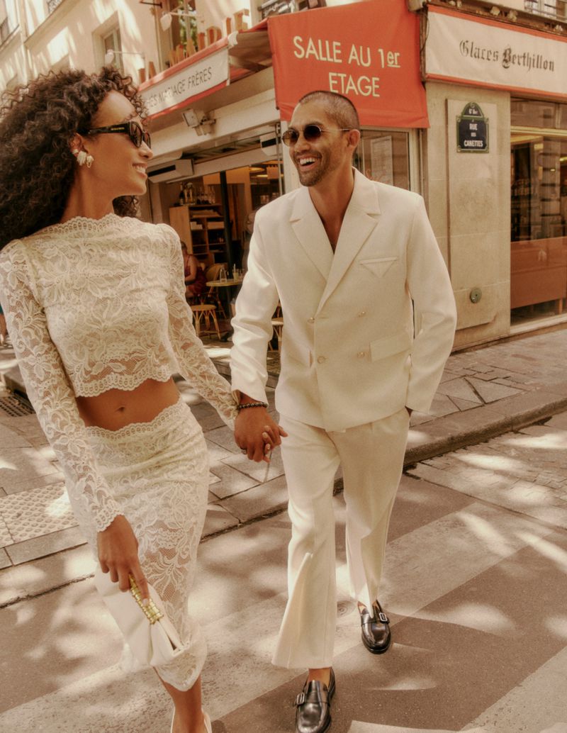 A stylish couple strolling hand-in-hand down a Parisian street in matching white outfits with natural lighting and a happy mood.