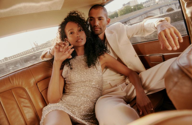 A stylish couple poses in the tan leather interior of a vintage car with a glimpse of a Parisian bridge and cityscape in the background.