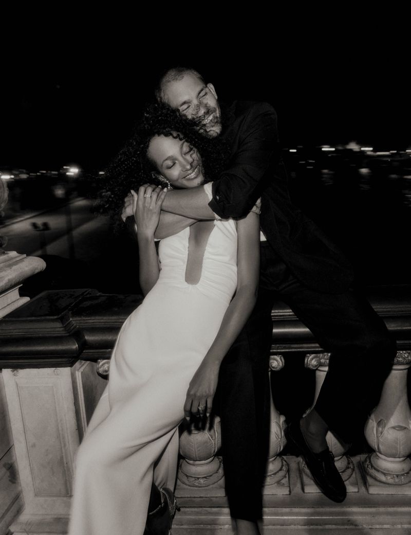 Romantic outdoor evening portrait of a smiling man embracing a woman in formal wear on a balcony overlooking a blurred city skyline.