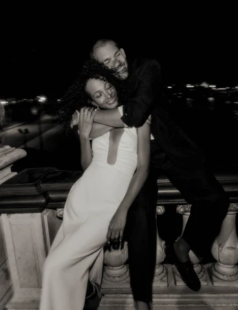 Romantic outdoor evening portrait of a smiling man embracing a woman in formal wear on a balcony overlooking a blurred city skyline.