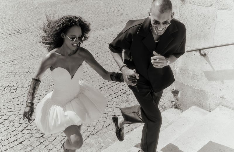 A fashionable couple is running up stone steps outside, with the woman in a short white dress and the man in a black suit.
